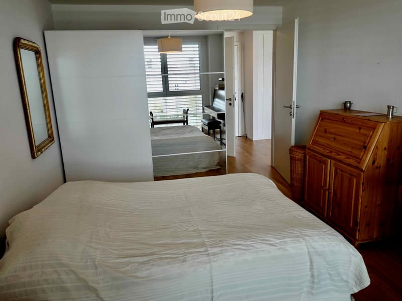 Spacious bedroom with a large bed, wooden dresser, mirror, and a view into an adjacent room with a chair and laptop.
