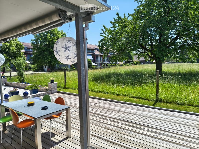 Wooden terrace with a dining table and colorful chairs, overlooking a green field and residential area.