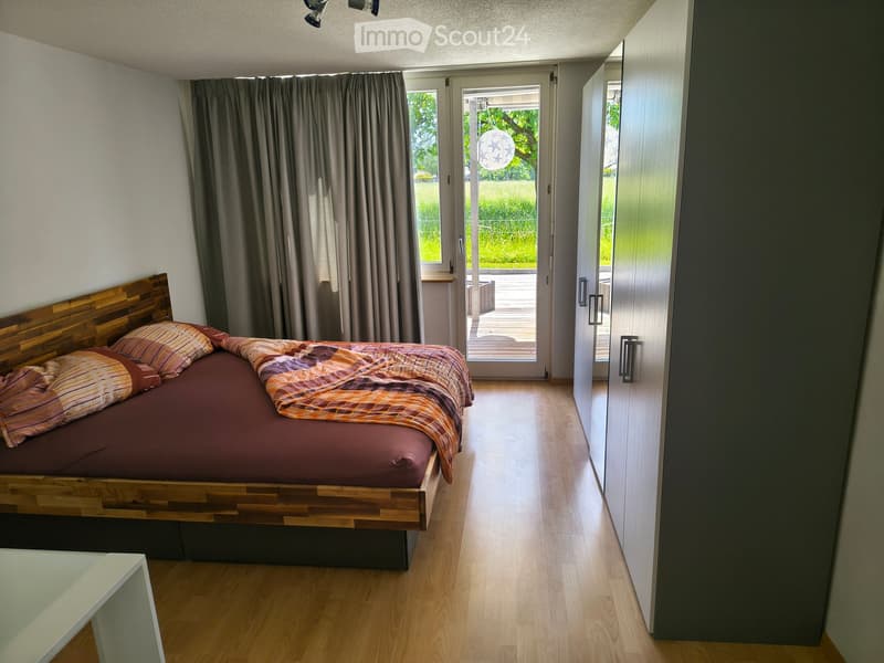 Modern bedroom with wooden bed frame, colorful bedding, nightstand, and large window with curtains leading to a balcony.