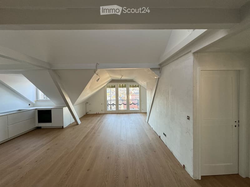 An attic space with wooden flooring, white walls, and large windows, featuring a small kitchen area with cabinets and a sink.