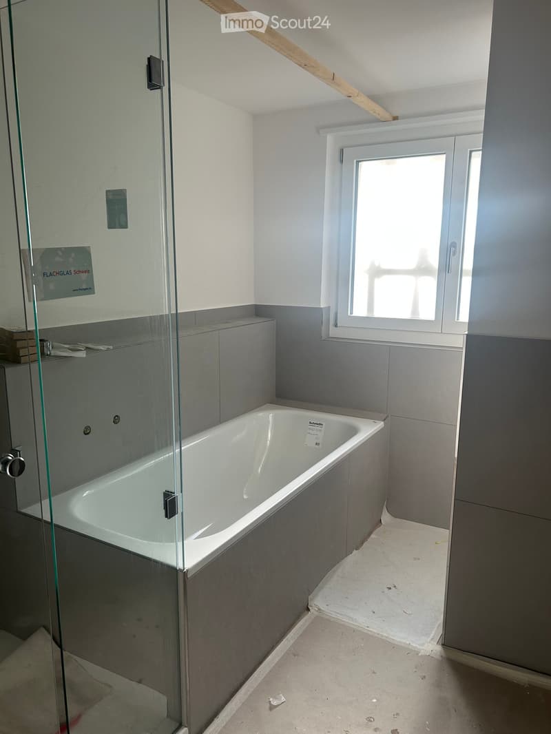 Modern bathroom with a white bathtub, glass shower enclosure, grey tiled walls, and a window providing natural light.