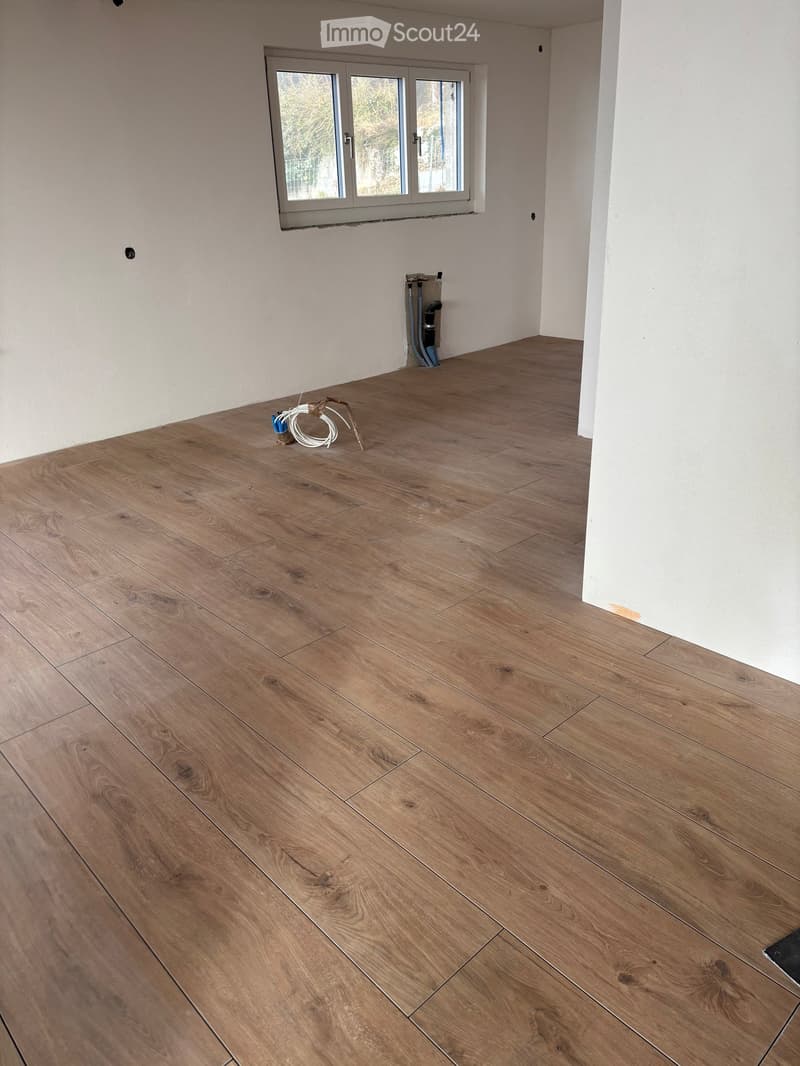 Empty room with wooden flooring and white walls, featuring a large window with a view of greenery outside.