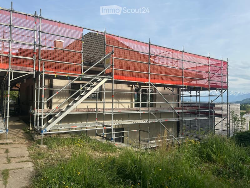 A two-story building under construction, covered with scaffolding and protective netting, with visible windows and a staircase leading to the upper level.