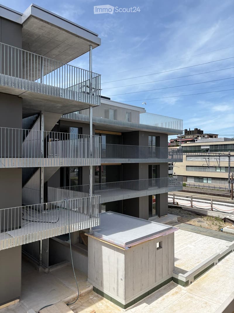 Modern multi-story building with balconies, metal railings, and a rooftop area featuring a small wooden structure and concrete surfaces.