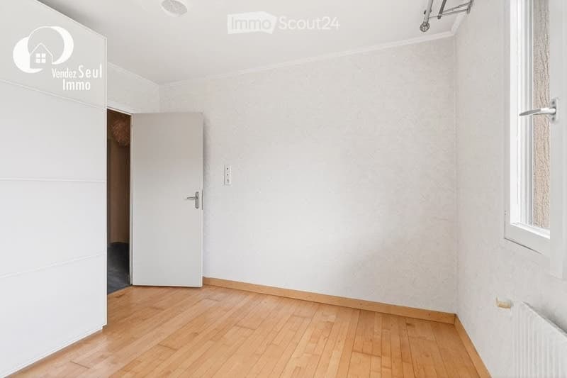 Empty room with light wooden flooring, white walls, and a single door leading to another space. The room has a window on one side and a radiator below it.