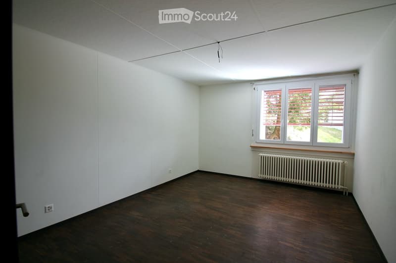 Empty room with white walls, dark wooden floor, and a single window with blinds. A radiator is placed beneath the window.
