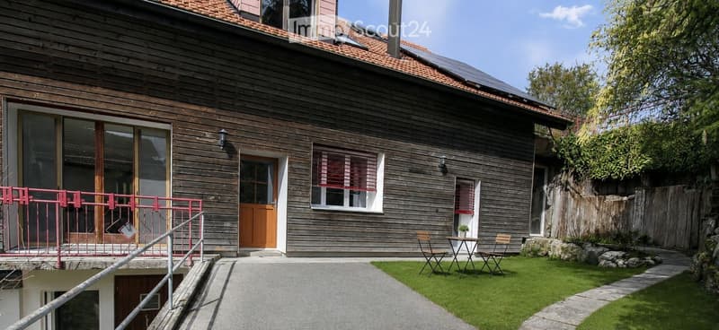 Two-story wooden house with red-tiled roof, large windows, and a small balcony area with a table and chairs.