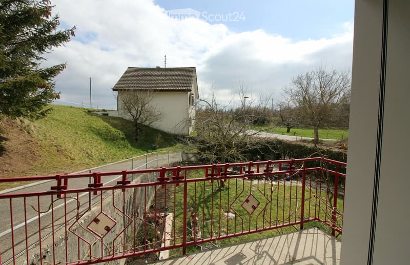 Balcony with red metal railing overlooking a hillside with a small house, trees, and a road below.