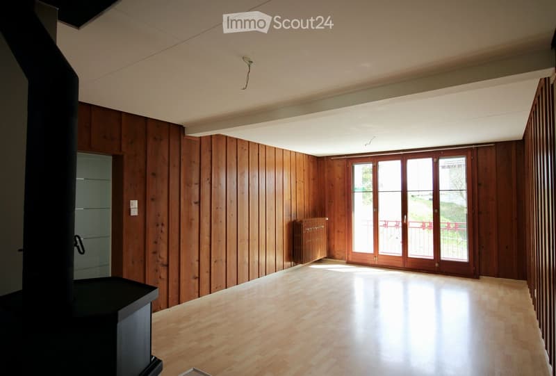 Empty room with wooden walls and floor, large windows with wooden frames, and a visible radiator. No furniture or equipment present.