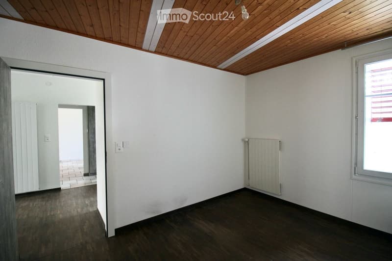 Empty room with wooden flooring, white walls, and a slanted wooden ceiling. Features a window with blinds, a radiator, and an open doorway leading to another space.