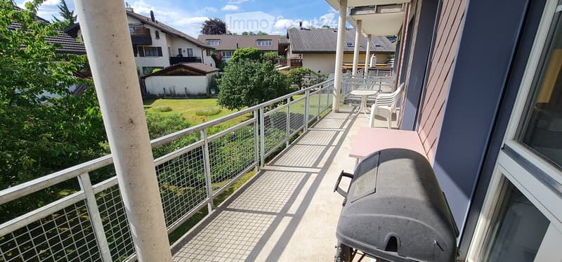 Balcony with metal railing, tiled floor, grill, table, and chairs, overlooking a green lawn and neighboring houses.