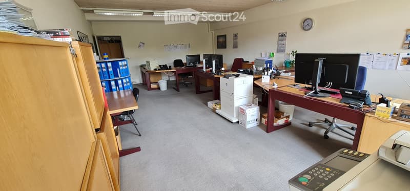 Office space with multiple desks, chairs, computer monitors, printers, and filing cabinets. The room includes a clock on the wall and a calendar.