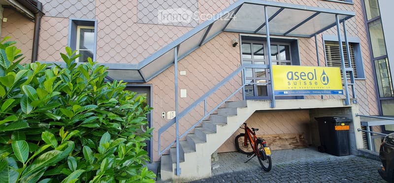 Exterior view of a modern building with a staircase leading to an entrance, a sign for "aseol suisse.ch", and a bicycle parked nearby.