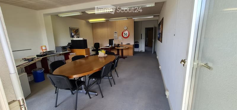 Office space with a round wooden table surrounded by chairs, desks with computers, office chairs, and a printer. The room has carpeted flooring, white walls, and a partially visible hallway leading to another area.