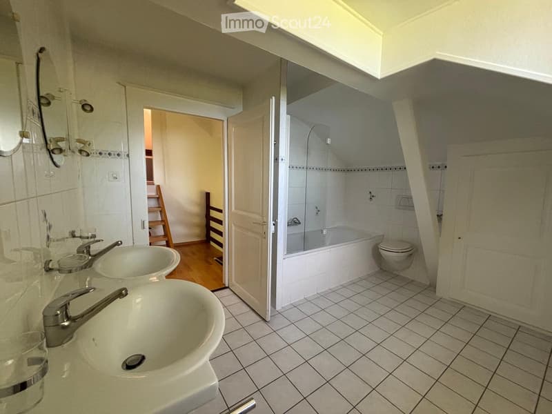 Bathroom with two sinks, a bathtub, and a toilet. The room has tiled floors and walls, with a wooden floor visible in the adjacent area.