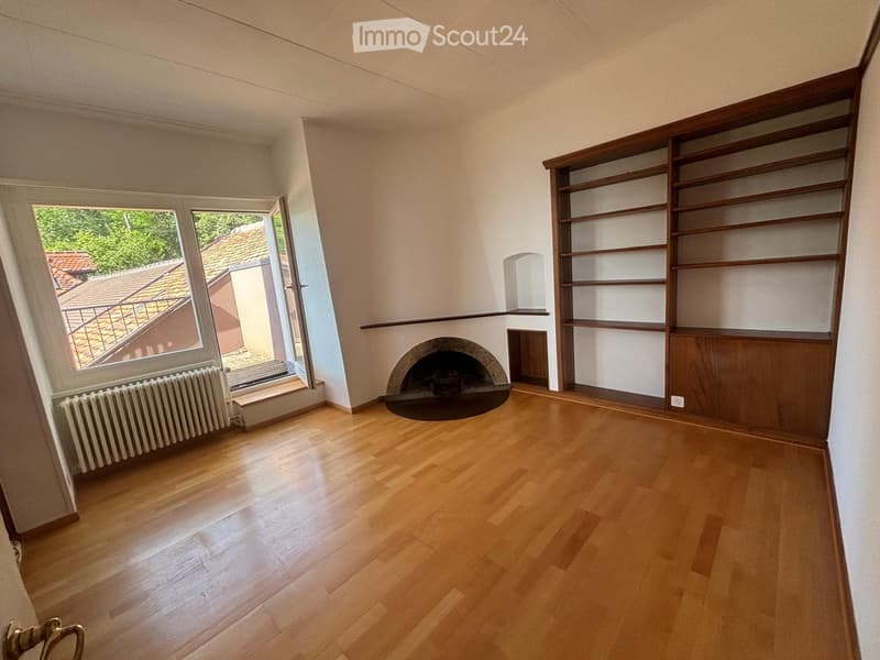 Empty room with wooden flooring, fireplace, built-in wooden shelving, large window with view of roof and greenery, radiator under window.