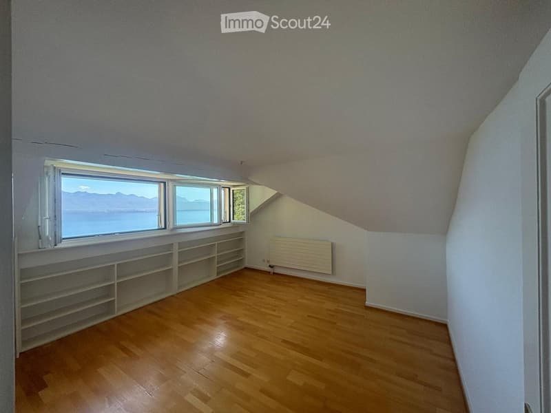 Bright, empty attic room with wooden flooring, built-in shelving, large windows offering a scenic view, and a radiator on one wall.