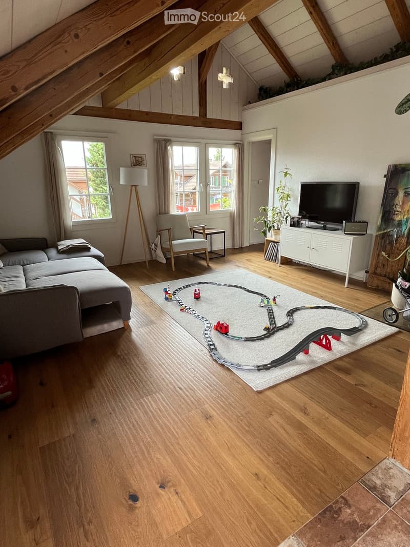 Bright living room with wooden floors, large windows, a grey sofa, armchair, coffee table, TV cabinet, floor lamp, and a toy train set on a white rug.