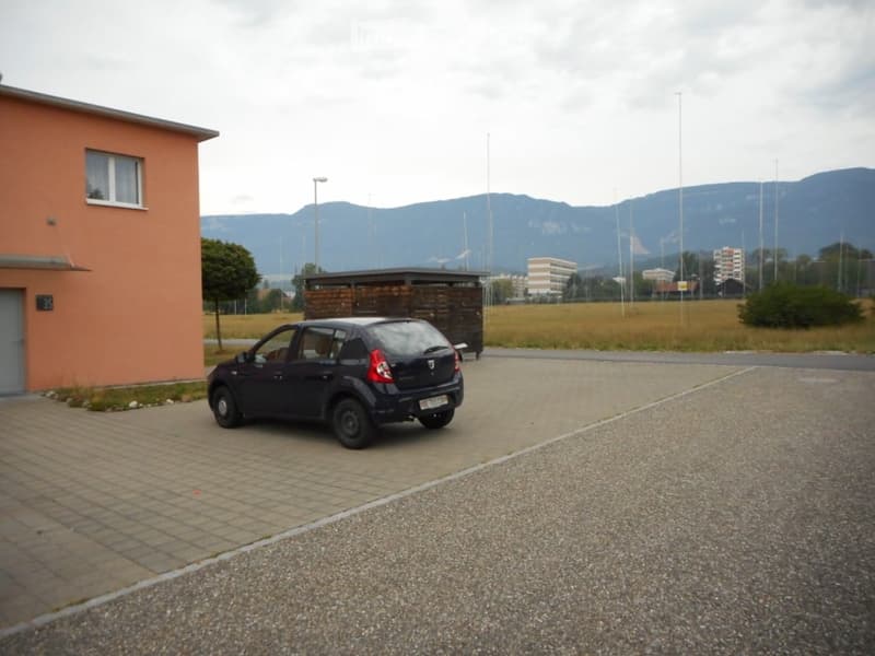 A small building with a parking area, a parked car, and a wooden shelter in the background, with a view of distant mountains.