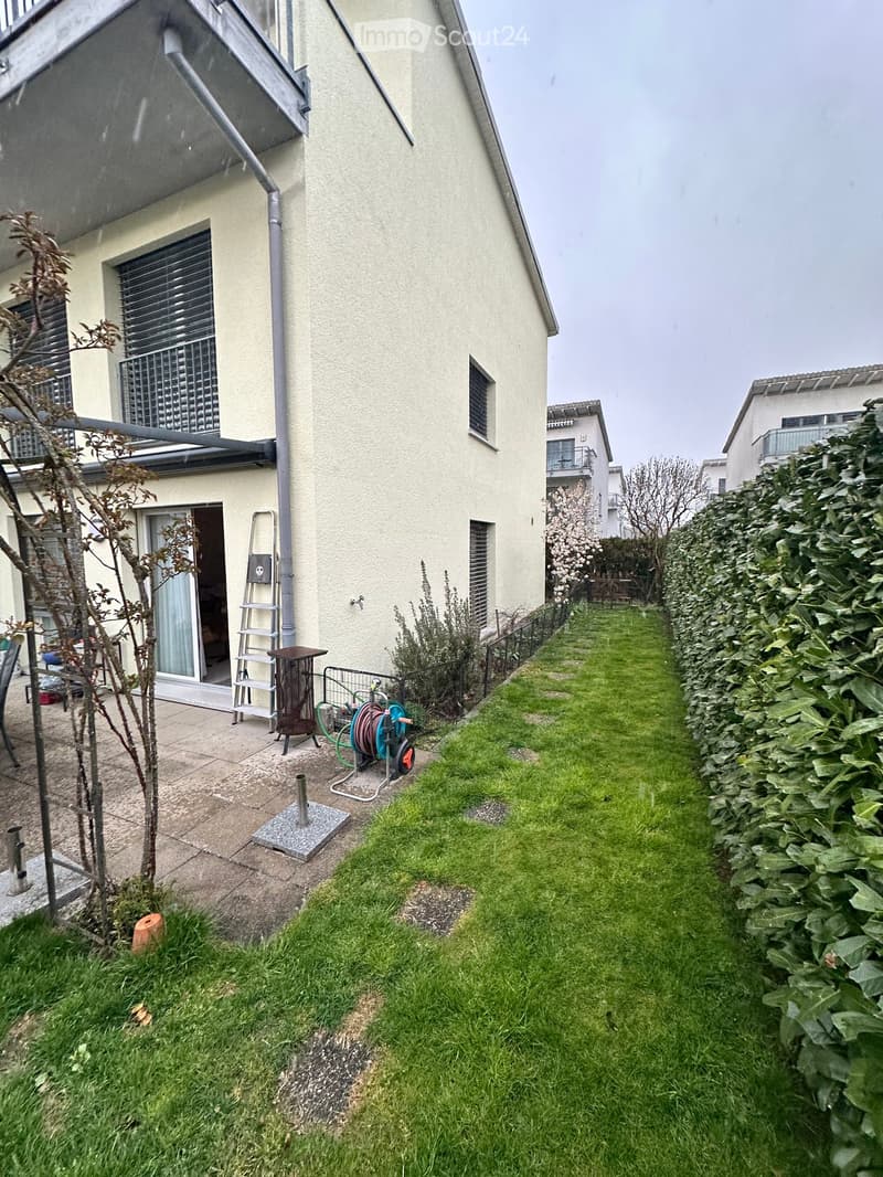 Garden area with a well-maintained lawn, a small patio with a ladder, a hose reel, and a small tree. The house has a light-colored exterior with a covered entrance and a window with blinds.