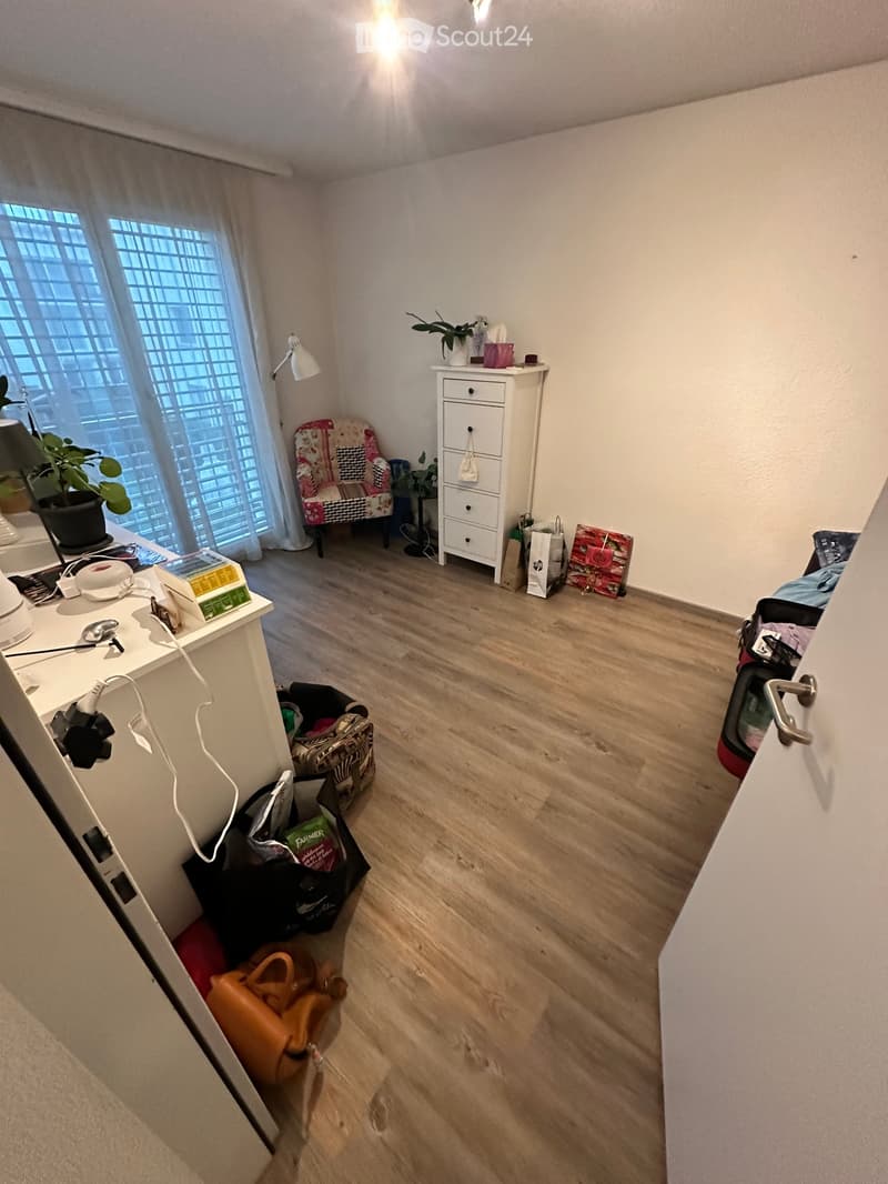 A small, furnished room with wooden flooring, a window with white curtains, a chair, a small cabinet, a table with a lamp, and several bags and items scattered around.