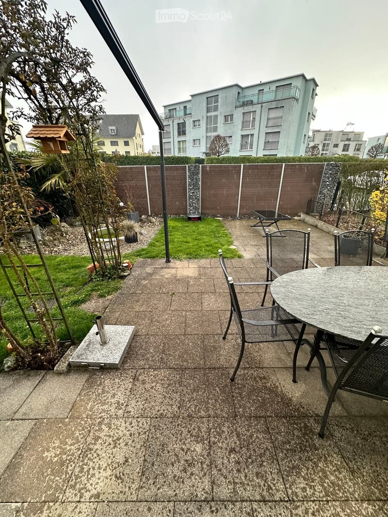 Outdoor patio with concrete floor, small table and chairs, small garden area with grass and plants, birdhouse, and modern apartment building in the background.