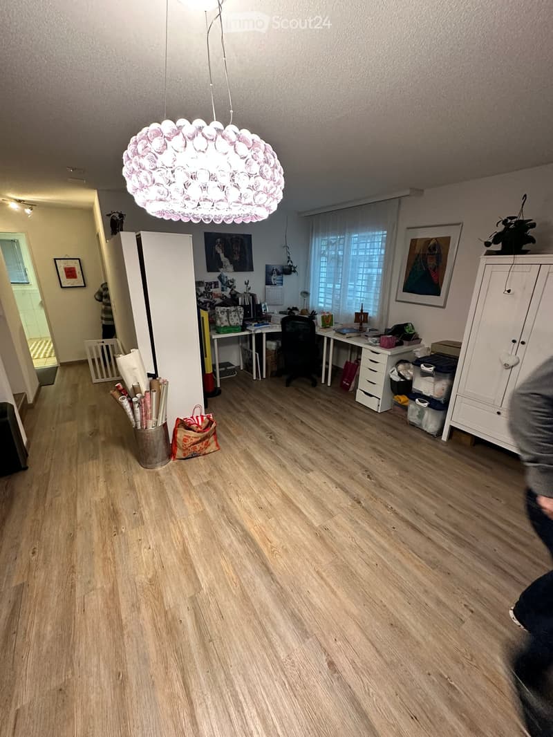 A well-lit room with wooden flooring, a large hanging lamp, a desk with a chair, a cabinet, and several picture frames on the walls.