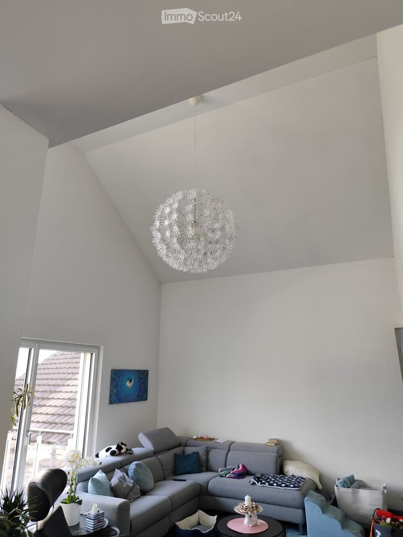 Modern living room with a grey sectional sofa, coffee table, side table, and decorative items. Large window with a view of a tiled roof, and a unique chandelier hanging from the sloped ceiling.