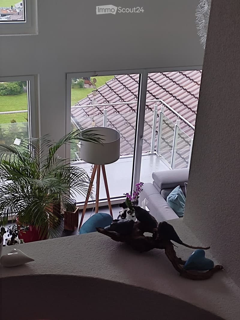 Bright living room with large windows and a balcony, featuring a sofa, a table with decorative items, a floor lamp, and potted plants.
