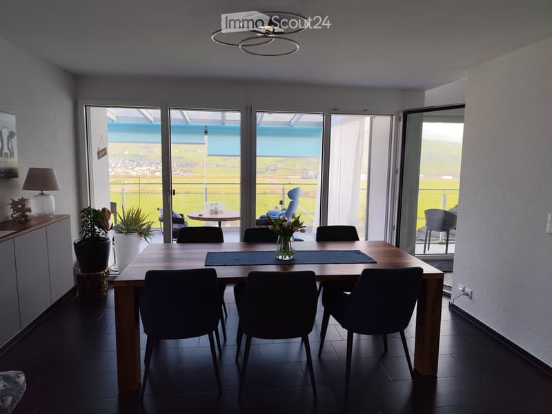 Modern dining room with a wooden dining table, six chairs, a vase with flowers, a table runner, a lamp, and a potted plant near the window. Large windows offer a scenic view of the landscape.