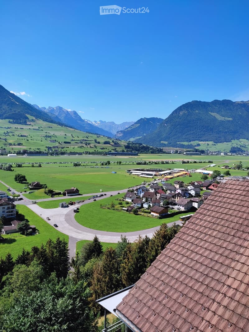 A panoramic view of a rural area with green fields, scattered houses, and distant mountains under a clear blue sky.