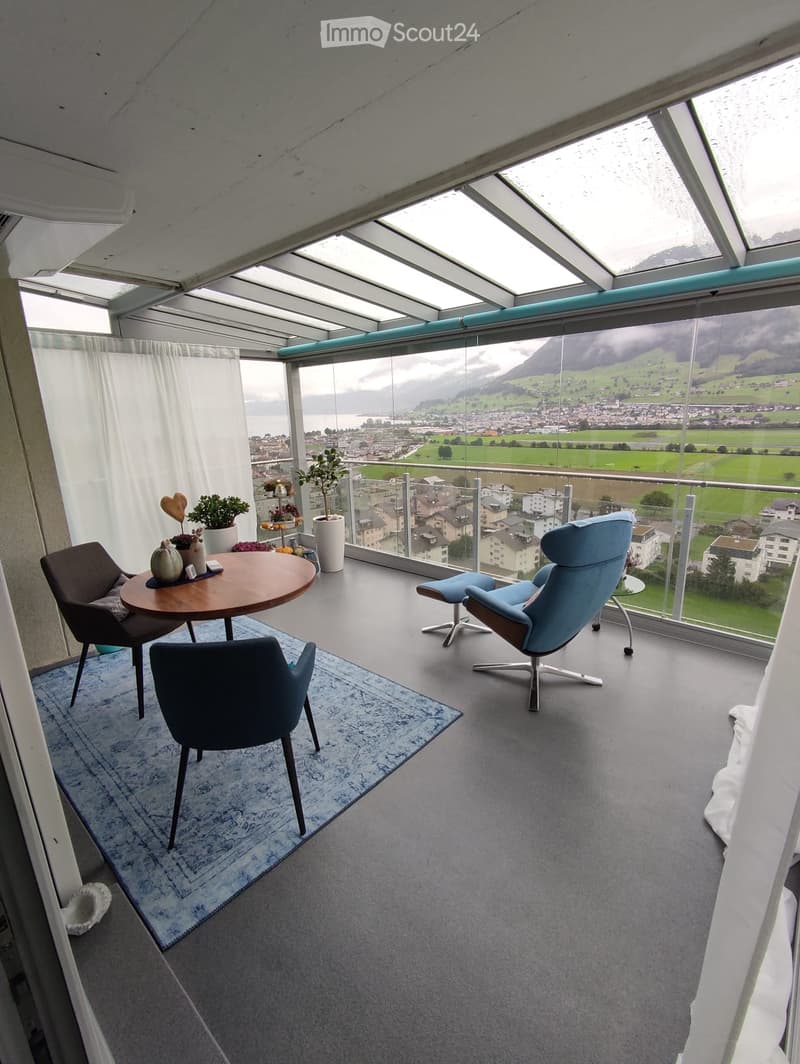 Modern balcony with panoramic mountain view, glass ceiling, grey carpet, blue rug, round wooden table, chairs, and potted plants.