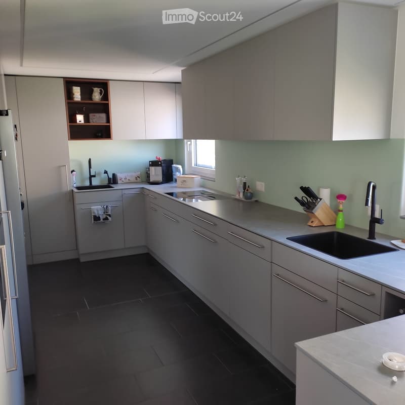 Modern kitchen with grey cabinets, dark tiled floor, and stainless steel sink. Includes a coffee maker, knife block, and a towel hanging from a cabinet.