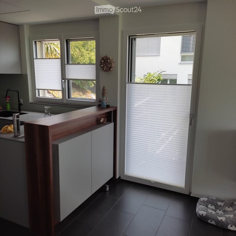 Modern kitchen with dark tiled floor, light grey walls, and large windows with blinds. Features a wooden counter, sink, faucet, and decorative items including a wreath and a small fountain figurine.