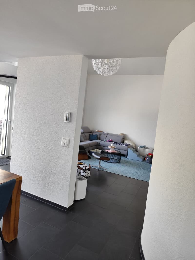 Modern living room with dark tiled flooring, white walls, and a grey sectional sofa. Features include a coffee table, a round side table, a hanging chandelier, and a small wooden dining table with a blue chair.