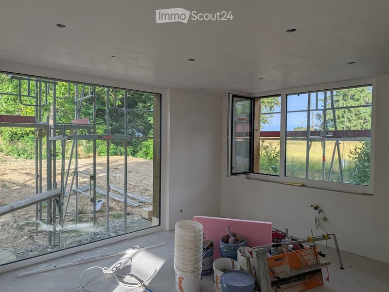 Empty room with bare walls and floor, large windows, and construction materials scattered around.