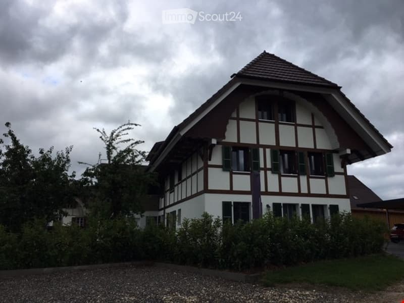 Traditional Swiss-style house with white walls, dark wooden trim, and a steeply pitched roof. The house features multiple windows and is surrounded by greenery and a gravel driveway.