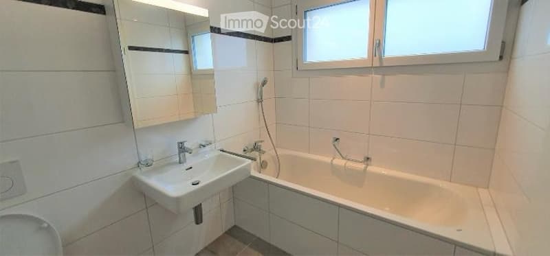 Modern bathroom with white tiles, a sink, a bathtub, and a large mirror above the sink. The room features a window for natural light and a sleek, contemporary design.