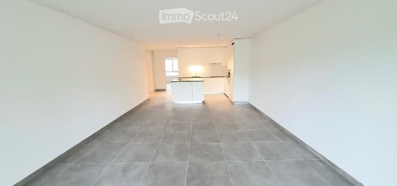 Empty room with white walls, grey tiled floor, and a small white kitchen area in the background.