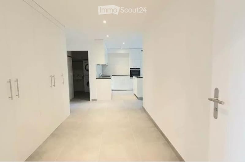 Bright, clean hallway with white walls, tiled floor, and built-in white cabinets on one side. The hallway leads to a kitchen area visible at the end.