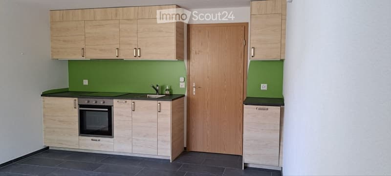 Modern kitchen with light wooden cabinets, green backsplash, black countertop, and built-in oven.