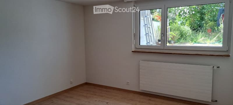 Empty room with wooden flooring, white walls, a radiator, and a large window with a view of greenery outside.