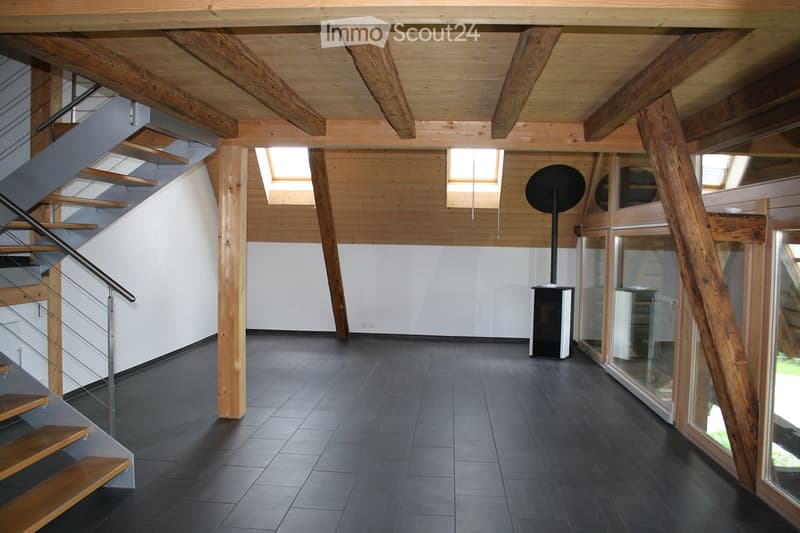 Open, modern interior with exposed wooden beams, dark tiled floor, staircase with wooden steps and metal railing, large windows, and a small black cylindrical object near the wall.