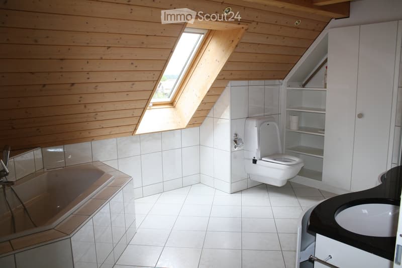 Modern bathroom with white tiled walls and floor, a toilet, a sink with a black countertop, a bathtub, and built-in white cabinets with shelves. The room features a skylight window and a wooden slanted ceiling.
