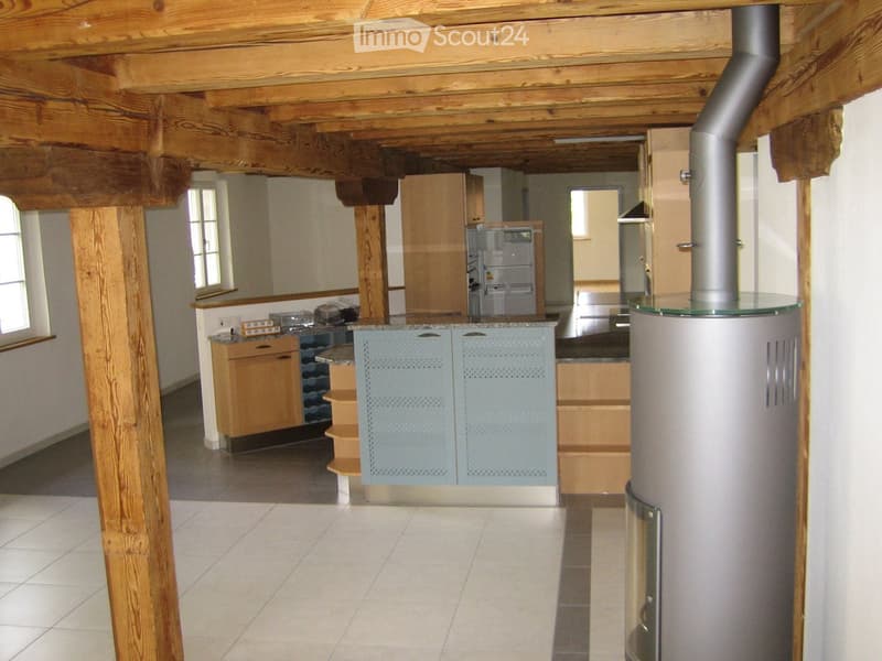 Open-plan kitchen and living area with exposed wooden beams, light-colored cabinets, a refrigerator, and a stove. The space features a tiled floor and large windows allowing natural light.