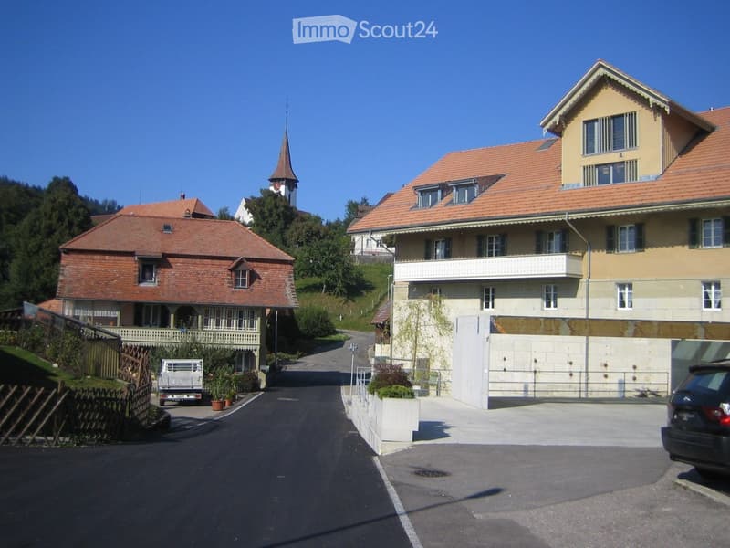 Two-story residential buildings with red-tiled roofs and white walls, featuring balconies and windows, set in a rural area with a church visible in the background.