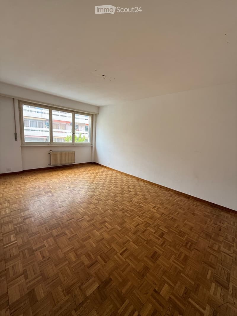 Empty room with wooden herringbone flooring, white walls, and large windows with a radiator below. No furniture or equipment present.