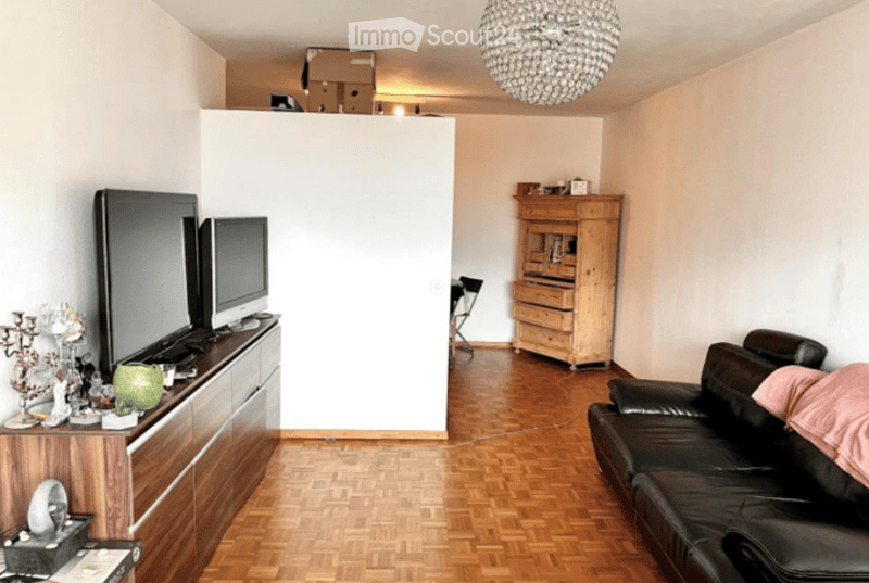 A modern living room with wooden flooring, a large white partition wall, a black leather sofa, a TV on a wooden stand, a wooden cabinet, and a chandelier hanging from the ceiling.