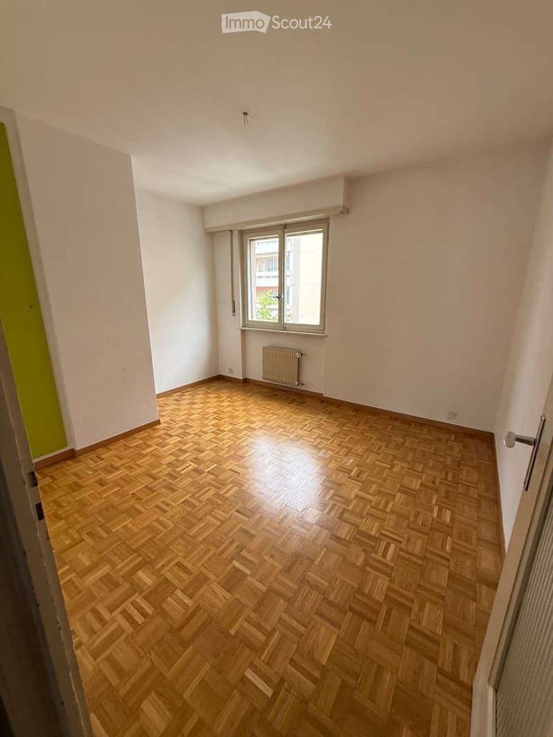 Empty room with wooden herringbone flooring, white walls, a window with blinds, and a radiator below it.