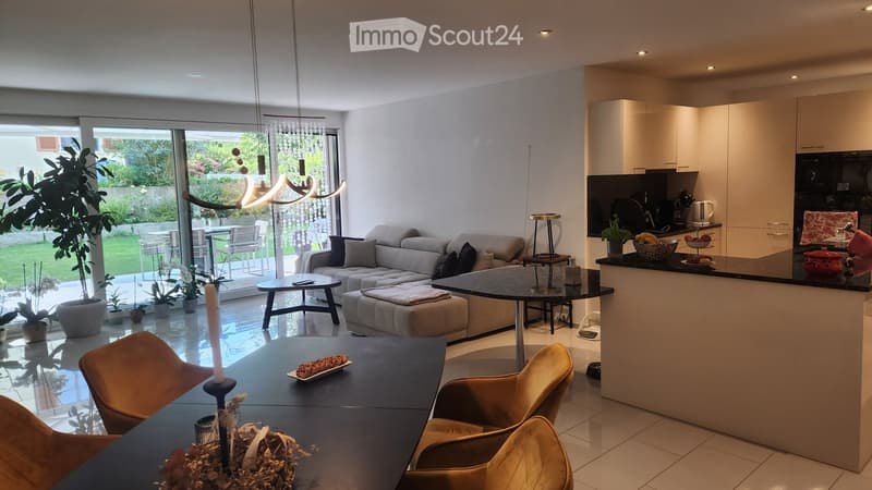 Open plan living room with dining area, couch, coffee table, kitchen island, dining table, plants, sliding glass doors, balcony, garden view